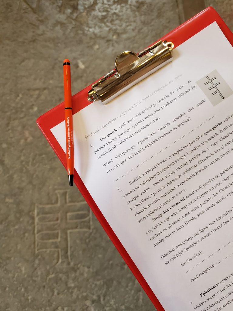 Zdjęcie w Centrum św. Jana. Zbliżenie na podkładkę piśmienniczą z kartą zadań dotyczących wyposażenia kościoła. W tle widoczna płyta nagrobna z gmerkiem kościoła św. Jana.